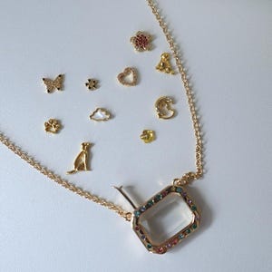 Peut inclure: Collier doré avec un pendentif rectangulaire bordé de pierres précieuses colorées. La chaîne porte divers charms, dont un papillon, une fleur, un cœur, un chien, une empreinte de patte, un nuage, une lune et une petite fleur.