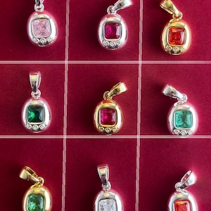 Puede incluir: Una colección de nueve colgantes ovalados dispuestos en una cuadrícula. Cada colgante presenta una piedra preciosa central de corte cuadrado en varios colores, incluyendo rosa, rojo, naranja, verde y transparente, con tres pequeñas piedras redondas.