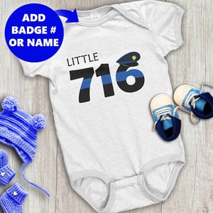 May include: White baby onesie with the text "LITTLE 716" and a police hat graphic. The image includes a blue knit hat, matching mittens, and blue and white baby shoes. The text "ADD BADGE # OR NAME" is in a blue circle.