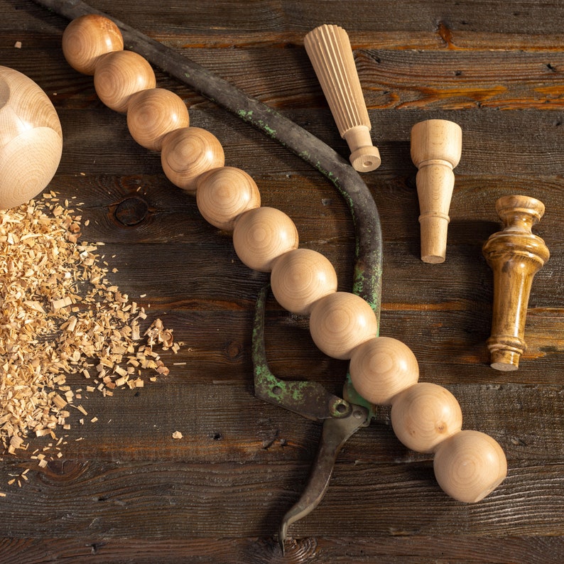 May include: A wooden dowel rod with a series of round wooden beads, a pair of wood turning tools, and a pile of wood shavings on a dark wood surface.