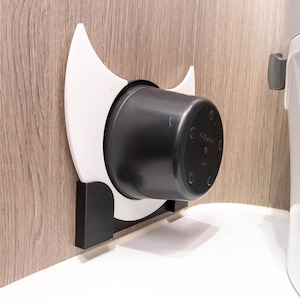 Könnte beinhalten: Ein weißer, wandmontierter Halter aus Kunststoff mit einem schwarzen Topf aus Kunststoff im Inneren. Der Halter ist halbmondförmig und hat eine schwarze Halterung aus Kunststoff an der Seite.