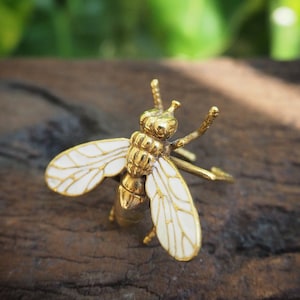 Könnte beinhalten: Eine goldfarbene Bienenbrosche mit weißen Emailleflügeln. Die Biene sitzt auf einem Stück Holz.
