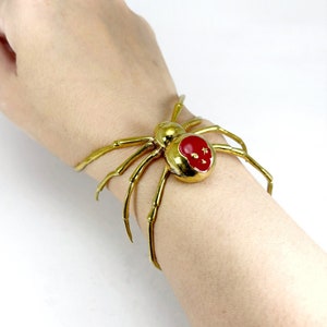 Puede incluir: Un brazalete de araña dorado con un centro rojo. El cuerpo de la araña es una gema redonda roja con detalles dorados. El brazalete se lleva en una muñeca sobre un fondo blanco.
