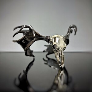May include: A silver deer skull with antlers, the skull is polished and shiny, the antlers are dark and textured, the skull is sitting on a black surface.
