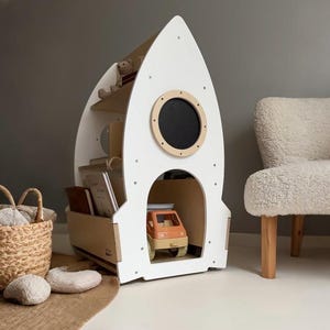 May include: White wooden rocket-shaped bookshelf with three shelves and a round porthole. The bookshelf is filled with books and a toy car.