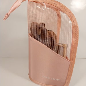 Puede incluir: Una bolsa de maquillaje de viaje rosa con un compartimento de malla y un cierre de cremallera. La bolsa tiene las palabras "TRAVEL SEASON" impresas en la parte delantera.