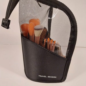 May include: Black mesh travel makeup bag with a zipper closure and a black fabric flap with the text "TRAVEL SEASON" printed in white. The bag is filled with makeup brushes and other makeup tools.