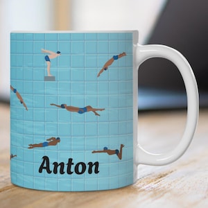 Könnte beinhalten: Weiße Keramik-Tasse mit hellblauem Hintergrund, auf dem Illustrationen von Schwimmern zu sehen sind, die in ein Schwimmbecken springen. Der Name "Anton" ist in Schwarz auf die Vorderseite der Tasse gedruckt. Die Tasse steht auf einer Holzoberfläche.