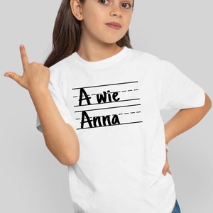 May include: White t-shirt with black text that says "A wie Anna" on it. The text is written in a handwritten style.