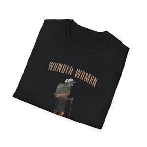 Puede incluir: Camiseta negra con el texto "WANDER WOMAN" en letras marrones. Una silueta de una persona haciendo senderismo con una mochila se superpone al texto.