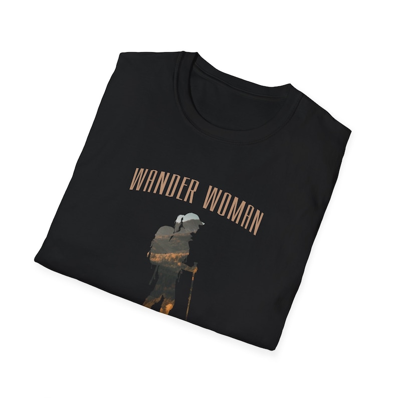 Puede incluir: Camiseta negra con el texto "WANDER WOMAN" en letras marrones. Una silueta de una persona haciendo senderismo con una mochila se superpone al texto.