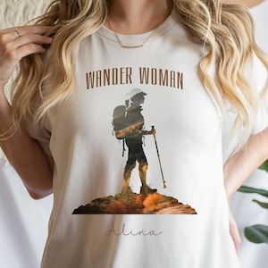 Puede incluir: Camiseta blanca con una silueta marr&oacute;n de una persona haciendo senderismo con una mochila y un bast&oacute;n de senderismo. El texto "Wander Woman" est&aacute; por encima de la silueta y el nombre "Alina" est&aacute; por debajo de la silueta.