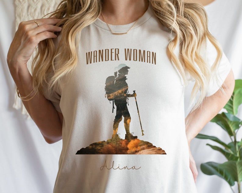 Puede incluir: Camiseta blanca con una silueta marr&oacute;n de una persona haciendo senderismo con una mochila y un bast&oacute;n de senderismo. El texto "Wander Woman" est&aacute; por encima de la silueta y el nombre "Alina" est&aacute; por debajo de la silueta.