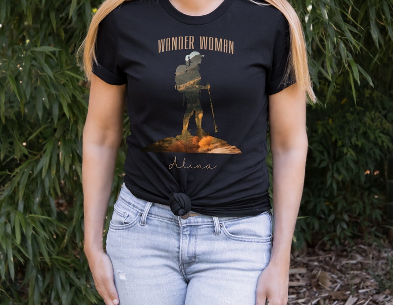 Puede incluir: Camiseta negra con un gr&aacute;fico de una excursionista con una mochila y bastones de senderismo. El texto "Wander Woman" est&aacute; por encima del gr&aacute;fico y el nombre "Alina" est&aacute; por debajo del gr&aacute;fico.