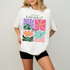 Könnte beinhalten: Weißes T-Shirt mit einem farbenfrohen Blumendesign. Das Design zeigt vier quadratische Felder mit stilisierten Blumen in Lila, Rosa, Koralle und Türkis, mit gelben Zentren. Der Text "Offizielles Schlafshirt" und der Name "Alina" sind ebenfalls aufgedruckt.