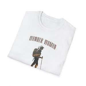 Puede incluir: Camiseta blanca con el texto "Wander Woman" impreso en marr&oacute;n. Debajo del texto se imprime una silueta de una persona con una mochila y un bast&oacute;n de senderismo.