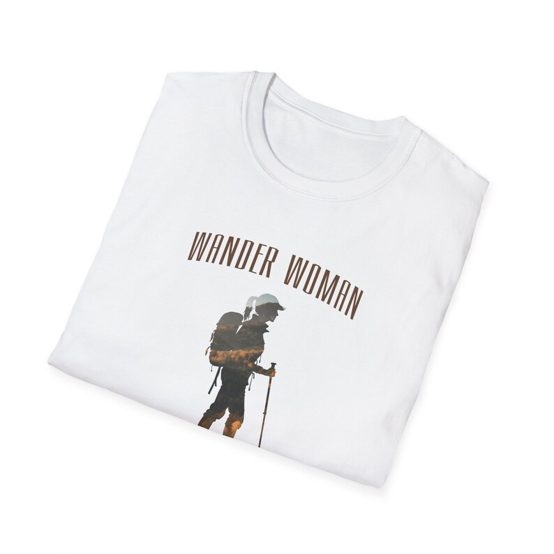 Puede incluir: Camiseta blanca con el texto "Wander Woman" impreso en marr&oacute;n. Debajo del texto se imprime una silueta de una persona con una mochila y un bast&oacute;n de senderismo.
