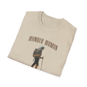 Puede incluir: Una camiseta beige con el texto "WANDER WOMAN" impreso en marr&oacute;n. La camiseta presenta la silueta de una persona haciendo senderismo con una mochila.