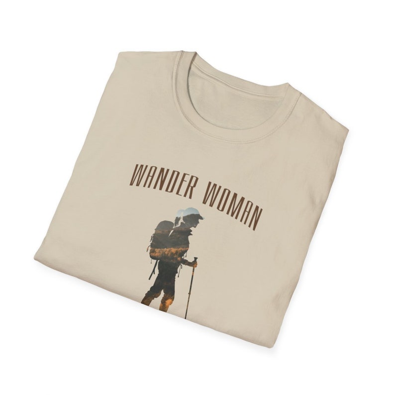 Puede incluir: Una camiseta beige con el texto "WANDER WOMAN" impreso en marr&oacute;n. La camiseta presenta la silueta de una persona haciendo senderismo con una mochila.