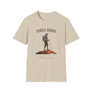 Puede incluir: Una camiseta beige con un gr&aacute;fico marr&oacute;n de una mujer haciendo senderismo con una mochila y el texto "Wander Woman" sobre el gr&aacute;fico y "Alina" debajo del gr&aacute;fico.