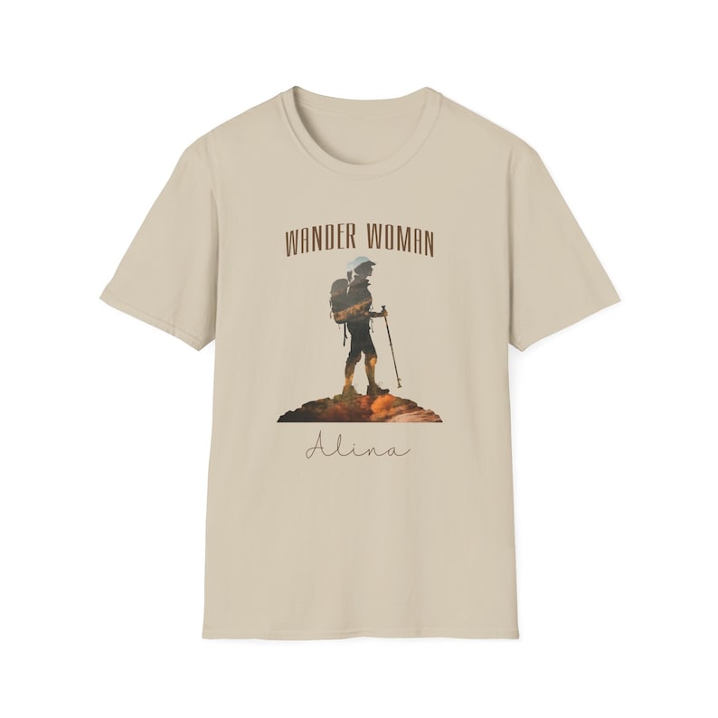 Puede incluir: Una camiseta beige con un gr&aacute;fico marr&oacute;n de una mujer haciendo senderismo con una mochila y el texto "Wander Woman" sobre el gr&aacute;fico y "Alina" debajo del gr&aacute;fico.