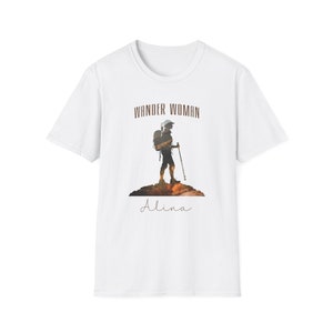Puede incluir: Camiseta blanca con una silueta marr&oacute;n de una persona haciendo senderismo con una mochila y bastones de senderismo. El texto "Wander Woman" est&aacute; por encima de la silueta y el nombre "Alina" est&aacute; por debajo de la silueta.