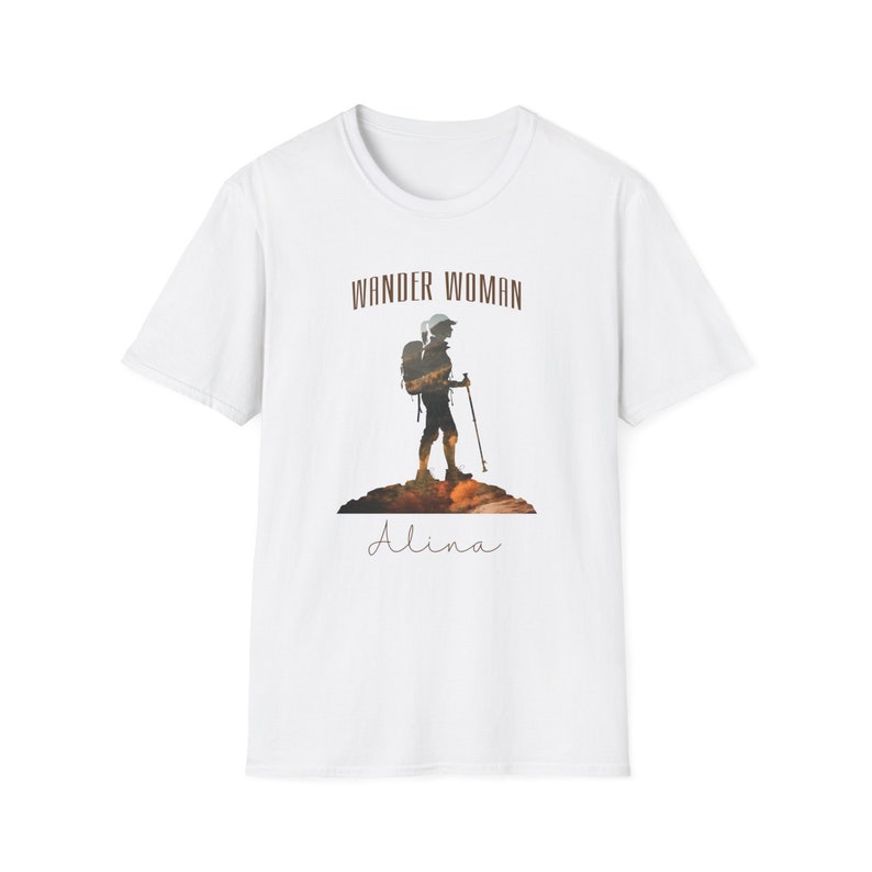 Puede incluir: Camiseta blanca con una silueta marr&oacute;n de una persona haciendo senderismo con una mochila y bastones de senderismo. El texto "Wander Woman" est&aacute; por encima de la silueta y el nombre "Alina" est&aacute; por debajo de la silueta.
