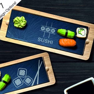 May include: A wooden serving tray with a black slate insert. The slate has a design of a noughts and crosses board with the word "SUSHI" underneath. The tray also has a small dish of wasabi and a few pieces of sushi on it.