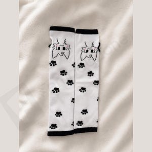May include: A pair of white arm warmers featuring a black cat face graphic at the top and a repeating pattern of small black paw prints throughout. Each arm warmer is finished with a solid black band at the top and bottom edges for a clean look.