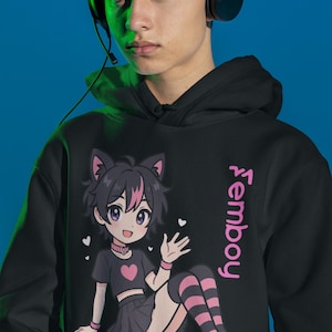 Op de afbeelding: Een zwarte hoodie met een grafische print van een anime-figuur met kattenoren, gekleed in een zwarte crop top met een roze hart, een geplooide rok en gestreepte kniekousen, met de tekst Femboy verticaal in het roze op de voorzijde gedrukt.