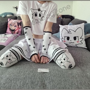 Puede incluir: Una camiseta corta blanca con un gráfico de gato negro, mangas de brazo blancas a juego con huellas de patas negras y una falda plisada blanca. El conjunto incluye calcetines blancos hasta la rodilla con una cara de gato y el texto BOYKISSER.