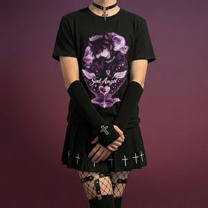 Pode incluir: Camiseta preta com um gráfico roxo de um personagem de anime e o texto "Sad Angel". O traje inclui uma saia plissada preta com cruzes brancas, aquecedores de braço pretos e um colar gargantilha.