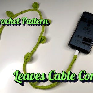 Può includere: Schema a uncinetto per un cavo a foglie verdi. Il cavo è avvolto attorno a un cavo di ricarica del telefono.