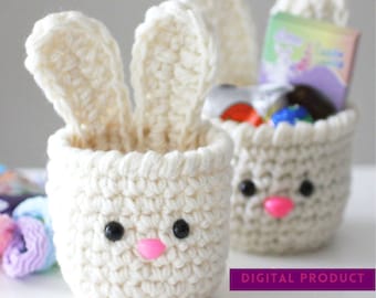 Può includere: Due cestini di coniglio in crochet bianco con occhi a bottone neri e nasi in feltro rosa. I cestini sono pieni di piccoli oggetti, tra cui una scatola viola e bianca con il testo "PRODOTTO DIGITALE".