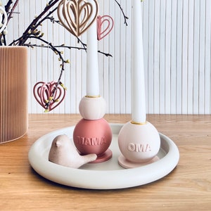 Könnte beinhalten: Zwei weiße Kerzenständer mit rosa und weißen Sockeln. Die Sockel sind mit den Worten "Mama" und "Oma" verziert. Die Kerzenständer stehen auf einem weißen Tablett mit einer kleinen Vogelfigur.