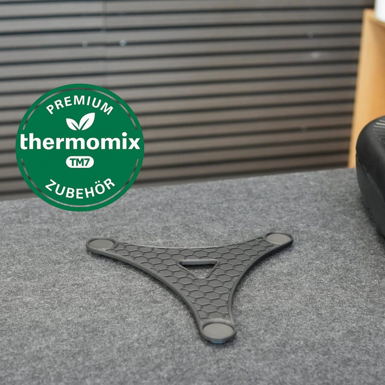 Op de afbeelding: Een donkergrijs, driehoekig keukenaccessoire met een honingraatpatroon en afgeronde randen. Het accessoire heeft een centrale driehoekige opening en is ontworpen voor gebruik met een Thermomix TM7. Een groen en wit cirkelvormig logo is zichtbaar.