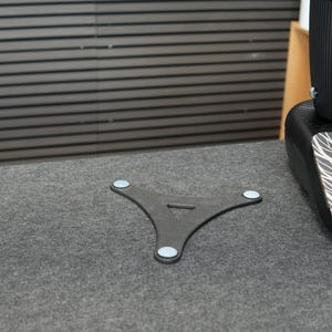 Op de afbeelding: Een donkergrijze, driepootstandaard met lichtblauwe pads op de voeten. De standaard staat op een grijs oppervlak, met een zwart object op de achtergrond. De standaard heeft een driehoekige vorm met een kleine opening in het midden.