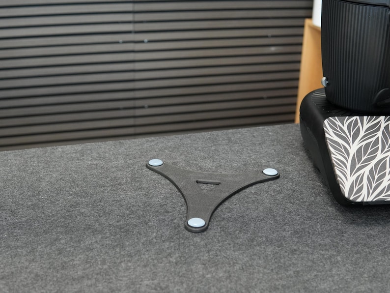Op de afbeelding: Een donkergrijze, driepootstandaard met lichtblauwe pads op de voeten. De standaard staat op een grijs oppervlak, met een zwart object op de achtergrond. De standaard heeft een driehoekige vorm met een kleine opening in het midden.