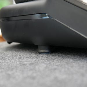 Op de afbeelding: Close-up van een zwart elektronisch apparaat met een glanzende bovenkant en matte zijkanten. Het apparaat rust op kleine, ronde voetjes. Het oppervlak eronder is een grijze, getextureerde stof. De afbeelding is goed belicht.