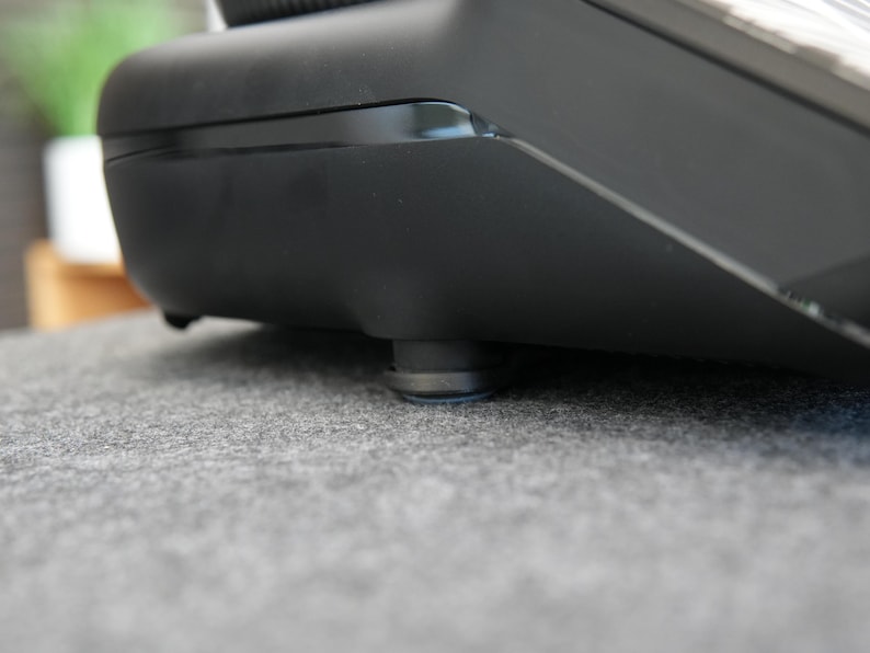 Op de afbeelding: Close-up van een zwart elektronisch apparaat met een glanzende bovenkant en matte zijkanten. Het apparaat rust op kleine, ronde voetjes. Het oppervlak eronder is een grijze, getextureerde stof. De afbeelding is goed belicht.