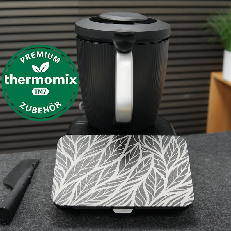 以下が含まれることがあります： シルバーのハンドルと蓋が付いた黒いキッチン家電。葉のデザインの模様が入った蓋が付いています。画像には、「Premium Thermomix TM7 Zubeh&ouml;r」と書かれた緑と白の円形ロゴが含まれています。
