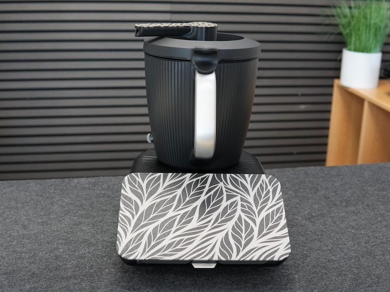 Op de afbeelding: Een zwarte elektrische waterkoker met een zilveren handvat en een patroon op het deksel. De basis heeft een bijpassend dienblad met een wit en grijs bladerenpatroon. De waterkoker staat op een donkergrijze ondergrond.