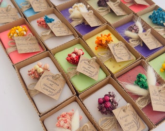 May include: A collection of rectangular soaps in individual cardboard boxes, each topped with dried flowers and a small kraft paper tag tied with twine. The tags feature printed names and dates, including Kristina & Manuel and Ivonne & Michael.