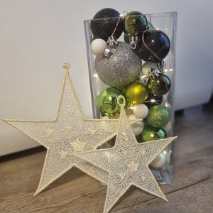 Può includere: Un vaso di vetro trasparente pieno di decorazioni natalizie verdi, argentate, nere e bianche. Due decorazioni a forma di stella traforata in primo piano. Le decorazioni sono su una superficie di legno.