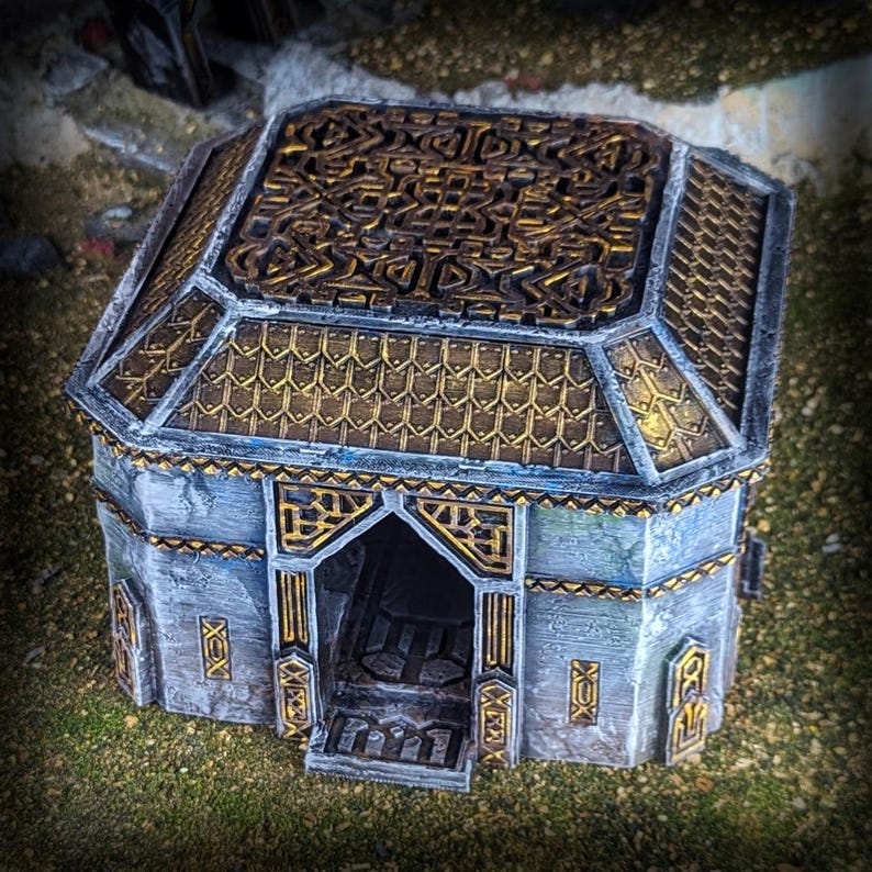 May include: A miniature fantasy building with a detailed design. The structure features a gray exterior with gold accents on the roof and trim. The roof has an intricate pattern, and the entrance is open, revealing interior details. The building is set on a grassy surface.