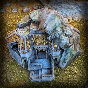 May include: A detailed miniature fantasy structure, resembling a dwarven stronghold or mountain hall. The model features a stone facade with gold accents, a rocky mountain top, and a detailed roof design. The overall color scheme is gray, gold, and brown.