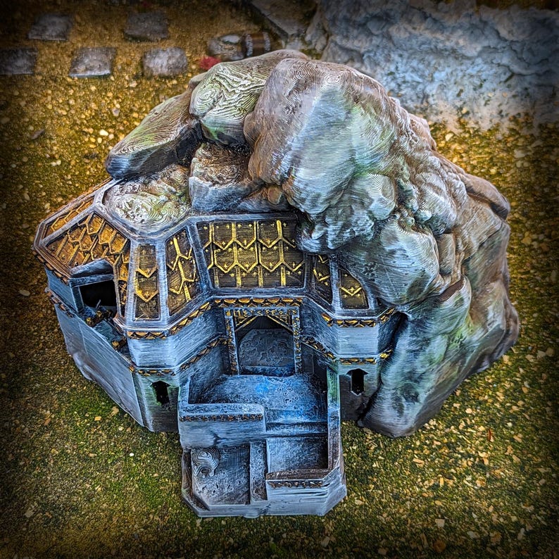 May include: A detailed miniature fantasy structure, resembling a dwarven stronghold or mountain hall. The model features a stone facade with gold accents, a rocky mountain top, and a detailed roof design. The overall color scheme is gray, gold, and brown.