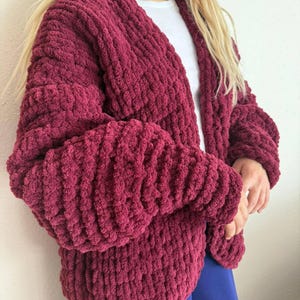 May include: A burgundy chunky knit cardigan with long sleeves. The open-front sweater is made of a thick, textured yarn. The sleeves are voluminous, and the cardigan appears soft and cozy. The person is wearing a white shirt underneath.
