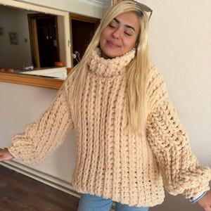 May include: A cream-coloured, chunky knit sweater with a high turtleneck collar and long sleeves. The sweater has a thick, textured appearance, with visible vertical ribbing. The sleeves have a slight bell shape at the cuffs. The sweater is made of a soft, plush material.