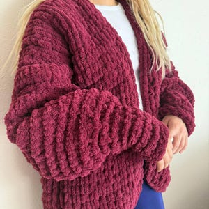 Puede incluir: Un cárdigan de punto grueso burdeos con mangas largas. El suéter abierto está hecho de un hilo grueso y texturizado. Las mangas son voluminosas y el cárdigan parece suave y cómodo. Se lleva una camiseta blanca debajo.